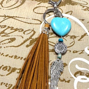 Puede incluir: Un llavero con un corazón turquesa, un dije de ala plateada y una borla marrón. El llavero tiene un cierre plateado y cuentas decorativas. El diseño incorpora elementos bohemios y del suroeste.