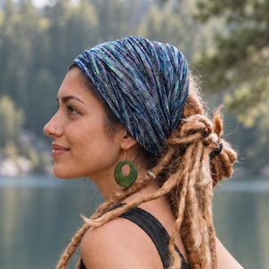 May include: A blue and purple tie-dye patterned head wrap worn as a headband, paired with a black sleeveless top and circular olive green earrings. The head wrap features a textured fabric with a mix of dark blue, light blue, and purple tones.