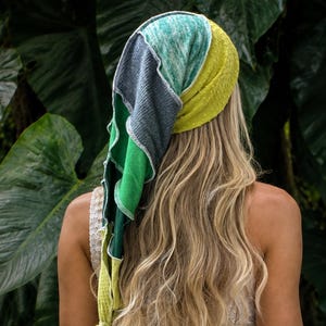 May include: A multi-coloured head scarf featuring patchwork panels in shades of teal, dark grey, bright green, and yellow with a textured finish. The scarf is tied at the back and worn over long blonde hair, paired with a white sleeveless lace dress.