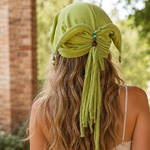 May include: A lime green fabric head covering featuring a gathered back with a central wooden bead accent and dangling fabric fringe. The head covering includes small decorative beads hanging from the side corners of the fabric piece.