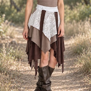 May include: A taupe tank top paired with a layered handkerchief skirt featuring brown fabric panels and a white lace overlay with a geometric pattern. The outfit is completed with dark brown leather knee-high boots with side buckle straps.