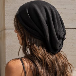 May include: A solid black slouchy beanie hat with a textured fabric finish. The headwear features a relaxed, gathered fit that sits loosely on the head, creating soft folds and a draped silhouette. The material has a subtle, fine-knit pattern.