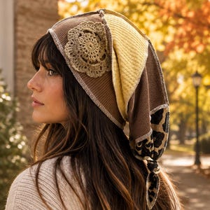 Puede incluir: Un pañuelo de retazos con tela acanalada marrón, un panel texturizado amarillo y una sección con estampado de leopardo. El pañuelo está decorado con una aplicación de flor de ganchillo beige y se ata en la parte posterior con costuras blancas.