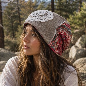 May include: A slouchy beanie hat featuring a heathered tan knit fabric, a white circular crochet lace appliqué on the top, and a vibrant red, pink, and teal patterned fabric panel along the back and side, creating a layered and textured appearance.