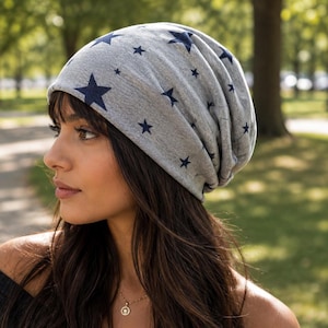 Może przedstawiać: Luźna czapka typu beanie z szarej melanżowej tkaniny z powtarzającym się wzorem małych, ciemnogranatowych gwiazdek. Czapka ma swobodny, nieusztywniony krój, który wygodnie układa się na głowie, z marszczonym wykończeniem z tyłu.