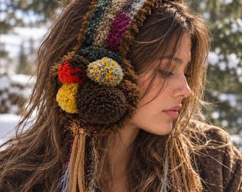 May include: A crochet ear warmer featuring a multi-colored striped headband in shades of green, brown, and magenta. The side displays a cluster of four yarn pom-poms in red, yellow, and brown, with long, dangling tan and light blue fringe tassels.