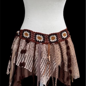 May include: A bohemian-style skirt with a crochet waistband featuring daisy-like patterns. The skirt is made of layered lace and fabric in shades of brown and beige, with dangling tassels and pom-poms. The skirt is displayed on a white mannequin.