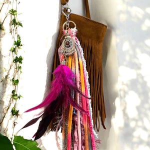 Puede incluir: Un llavero colorido de estilo bohemio con un cierre plateado, que presenta un dije decorativo, cintas vibrantes y un grupo de plumas rosas y moradas. Una bolsa de gamuza marrón con flecos cuelga en el fondo.