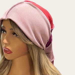 May include: A multi-colored head wrap featuring a wide, ribbed light pink front band. The back portion of the head wrap transitions into solid panels of bright red and deep purple fabric, constructed with visible seams along the top and sides.