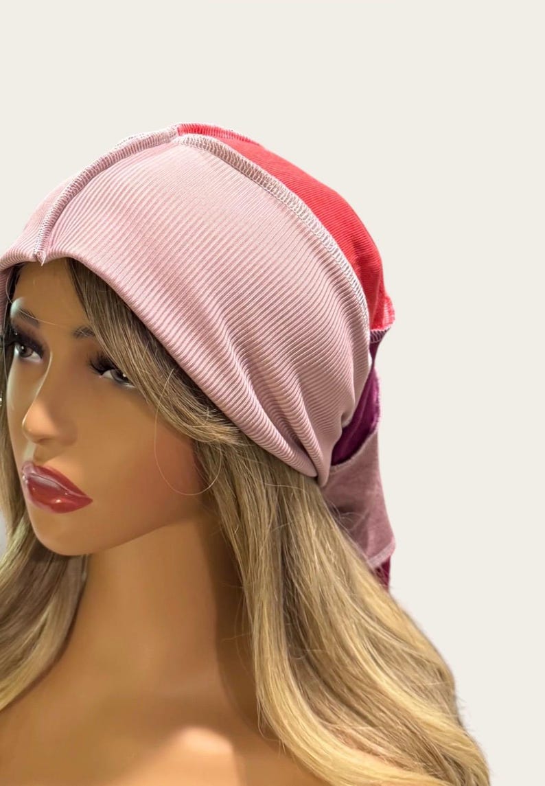 May include: A multi-colored head wrap featuring a wide, ribbed light pink front band. The back portion of the head wrap transitions into solid panels of bright red and deep purple fabric, constructed with visible seams along the top and sides.