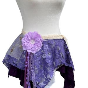 Puede incluir: Una falda morada en capas con un gran acento floral lavanda. La falda presenta encaje y telas transparentes, con un ribete decorativo y cintas colgantes. La falda se exhibe en un maniquí blanco.