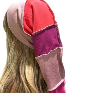 May include: A long, tubular head covering constructed from multiple fabric panels in shades of light pink, bright red, deep purple, mauve, and magenta. The panels are joined with visible, light-colored overlock stitching along the seams.
