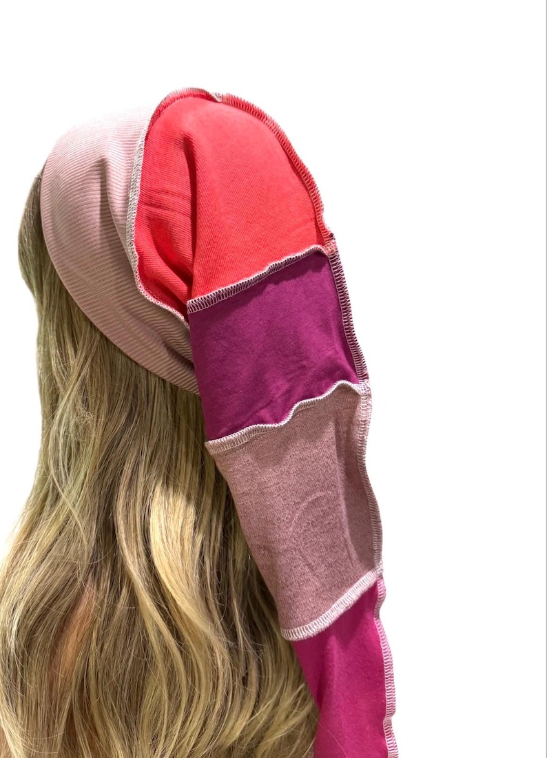 May include: A long, tubular head covering constructed from multiple fabric panels in shades of light pink, bright red, deep purple, mauve, and magenta. The panels are joined with visible, light-colored overlock stitching along the seams.