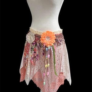 May include: A bohemian-style skirt in shades of peach, cream, and floral prints. The skirt features lace, ribbons, and a large orange flower. Colorful pom-poms and a green ribbon hang from the waist. The skirt is displayed on a white mannequin.