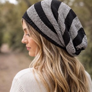 May include: A slouchy beanie hat featuring a knit pattern with alternating wide horizontal stripes in solid black and a textured heathered gray. The fabric has a ribbed appearance, and the hat is designed with a relaxed, loose fit that drapes.