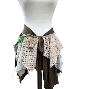 Puede incluir: Un envoltorio de cintura de estilo bohemio, exhibido en un maniquí. El envoltorio presenta capas de tela en verde oliva, crema y gris, con un adorno de flor de ganchillo. El diseño incluye detalles de encaje y cinta, creando un aspecto único y artesanal.