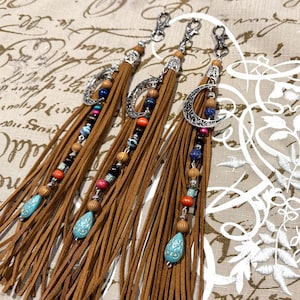 Puede incluir: Tres llaveros de borlas de gamuza marrón con herrajes plateados. Cada llavero presenta una cadena de cuentas decorativa con cuentas coloridas y un dije de lágrima turquesa. Un llavero tiene un dije de media luna.