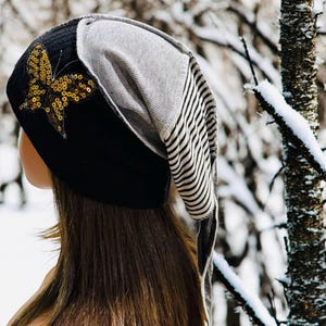 May include: A black beanie with a gold sequined butterfly applique. The hat has a gray and white striped section and a gray section. The hat is worn by a person with long brown hair, with a snowy forest in the background.
