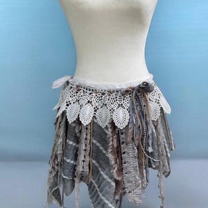 May include: A handmade, bohemian-style skirt with a layered design. The skirt features a white lace trim at the waist, with gray and brown fabric layers. Decorative elements include ribbons, beads, and lace, creating a unique, textured look.