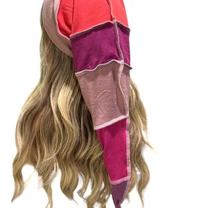 May include: A long, pointed beanie hat constructed from multiple fabric panels in shades of red, magenta, pink, and mauve. The hat features visible external seams with light-colored stitching, creating a patchwork design that tapers to a point.