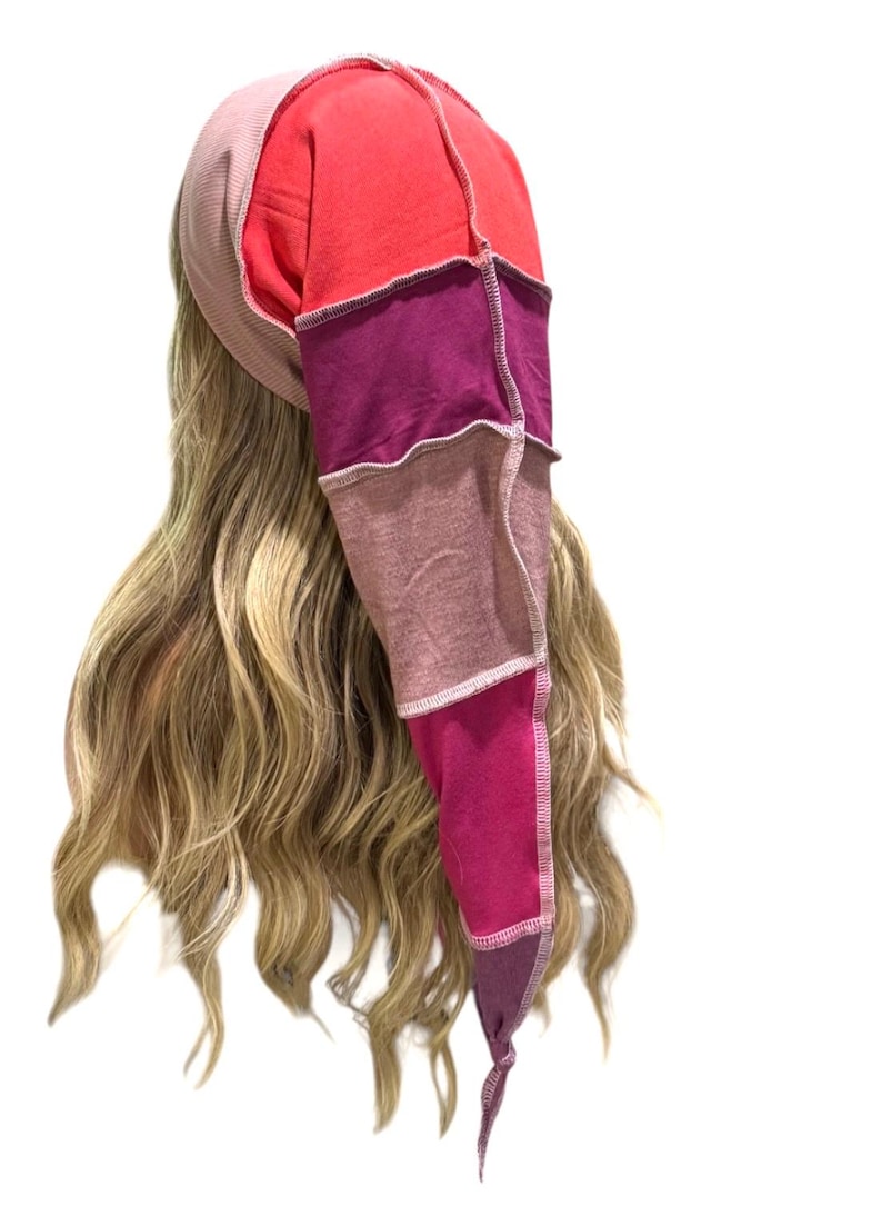 May include: A long, pointed beanie hat constructed from multiple fabric panels in shades of red, magenta, pink, and mauve. The hat features visible external seams with light-colored stitching, creating a patchwork design that tapers to a point.