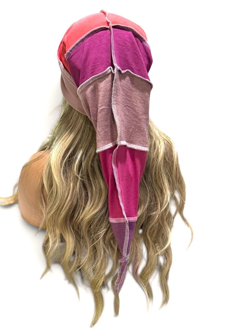 May include: A slouchy beanie hat constructed from multiple fabric panels in shades of red, magenta, purple, and dusty rose. The hat features prominent external overlock stitching in a light thread color, creating a patchwork design with a pointed tail.