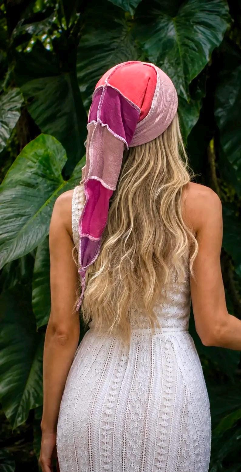 May include: A headscarf featuring a patchwork design of pink, magenta, and purple fabric panels with visible white stitching. The scarf has a long, pointed tail and is worn over long blonde hair, paired with a white sleeveless lace dress.