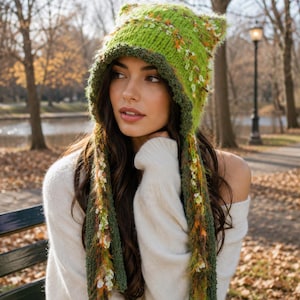 Puede incluir: Un gorro de punto verde lima con puntas en forma de orejas de gato y largos flecos laterales. El gorro presenta un borde acanalado verde oscuro y está adornado con acentos de hilo texturizado multicolor en tonos naranja, blanco y marrón.