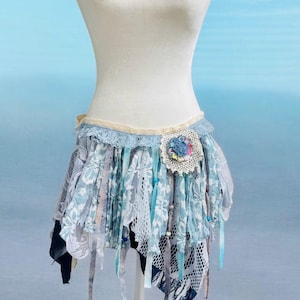 May include: A blue and white lace skirt with fabric strips and a floral decoration. The skirt is displayed on a mannequin. The skirt has a layered design with various textures and colours, including blue, white, and cream.