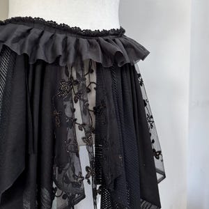 May include: Black skirt with a layered design, featuring a ruffled waistband and intricate floral lace panels. The skirt has a unique asymmetrical hemline and is displayed on a mannequin.
