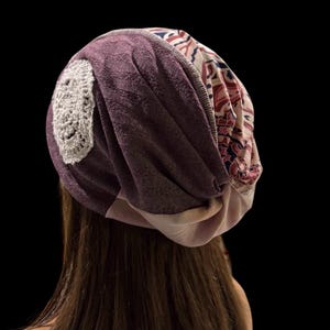 May include: A purple beanie with a crocheted patch and patterned fabric accents. The hat is made of soft material and has a relaxed fit. The beanie is shown from the back, with long brown hair visible.
