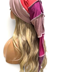 May include: A multi-colored head covering featuring a ribbed light pink front section, a bright red top panel, and a long, pointed back section constructed from patchwork fabric in shades of deep magenta, mauve, and dark purple with visible seams.