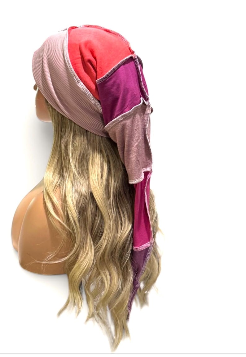 May include: A multi-colored head covering featuring a ribbed light pink front section, a bright red top panel, and a long, pointed back section constructed from patchwork fabric in shades of deep magenta, mauve, and dark purple with visible seams.