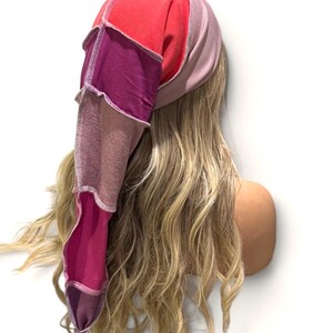 May include: A long, pointed beanie hat constructed from multiple fabric panels in shades of red, magenta, and purple. The hat features visible white overlock stitching along the seams, connecting the various colored sections into a tapered shape.