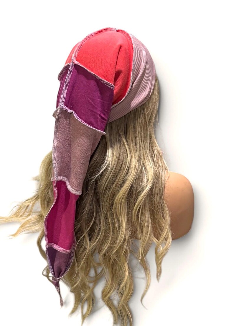 May include: A long, pointed beanie hat constructed from multiple fabric panels in shades of red, magenta, and purple. The hat features visible white overlock stitching along the seams, connecting the various colored sections into a tapered shape.