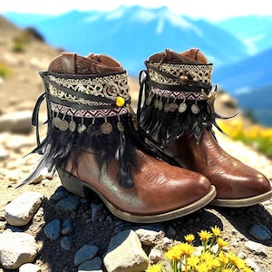 May include: A pair of brown leather ankle boots featuring decorative trim with gold embroidery, black leather straps, small dangling metallic coins, and black feathers. The boots have a rounded toe and a low heel, with a side zipper closure.