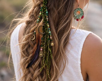 Puede incluir: Un accesorio para el cabello con hebras de lana verde, dijes de metal dorado con forma de sol y luna, y cuentas multicolores. La pieza incluye plumas marrones y negras unidas a las hebras, creando un diseño colgante y en capas.