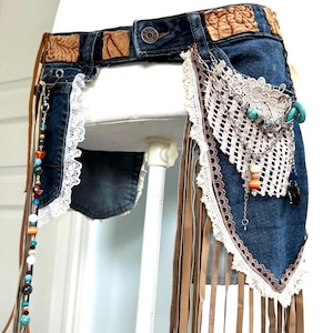 Könnte beinhalten: Ein Jeansgürtel mit dekorativen Elementen. Der Gürtel hat braune Lederpatches, weiße Spitzenbesätze und baumelnde Lederstreifen. Er ist mit Perlen, Türkissteinen und einer gehäkelten Tasche verziert.