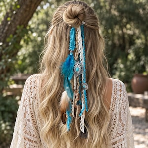 Könnte beinhalten: Ein Haar-Accessoire bestehend aus blauen und beigen Federn, türkisfarbenen Steinen in silberfarbenen Metallfassungen, weißer Spitze und blauen Stoffstreifen, befestigt an einer Spange in langem, halb hochgestecktem blondem Haar.