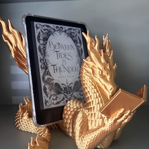 Op de afbeelding: Een metallic gouden drakenfiguur die een klein boek vasthoudt, ontworpen als standaard voor een e-reader. De e-reader toont de boekomslag met de titel Between Tides & Thunder van Leena Kazak, voorzien van gedetailleerde zwart-witte golfillustraties.