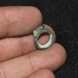 May include: A small, weathered, circular metal ring with a dark, oxidized grey-green patina and a textured, uneven surface. The band features a slightly raised, worn section on one side, showing signs of age and metallic corrosion throughout.