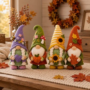 May include: Four handmade crochet gnomes standing in a row. The first wears a purple hat with flowers and a bee, holding a purple heart. The second has a green hat with a mushroom, holding a pumpkin. The third has a yellow striped hat with a sunflower, holding a sunflower. The fourth wears an orange hat with autumn leaves, holding a leaf.