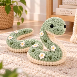 May include: A sage green and cream crochet snake plush toy featuring small white daisy appliques with yellow centres. The snake has two black plastic eyes and a coiled body shape. The texture is made of thick yarn loops in a soft, rounded design.