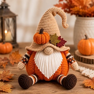 Può includere: Una statuetta di gnomo all'uncinetto con un cappello a punta color marrone chiaro decorato con una piccola zucca arancione e due foglie autunnali. Lo gnomo ha una barba di filo bianco, un corpo a forma di zucca arancione e piedi marroni.