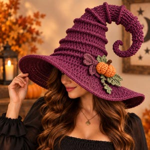 May include: A deep purple crocheted witch hat featuring a tall, pointed, and curved crown with a wide brim. The hat is decorated with a small, textured orange crocheted pumpkin and several green crocheted leaves attached to the side of the brim.