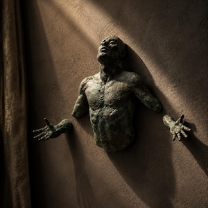 May include: A wall-mounted bronze-toned sculpture depicting a human torso with an upward-facing head and outstretched arms. The figure features a weathered, textured surface with dark green and brown patina, creating a dramatic effect.