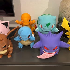 Könnte beinhalten: Eine Sammlung von sieben bunten Kunststoff-Figuren, darunter ein braunes Evoli, ein rosa Pummeluff, ein orangefarbenes Glumanda, ein blaues Schiggy, ein hellblaues Bisasam, ein violettes Gengar mit langer Zunge und ein gelbes Pikachu mit roten Wangen.