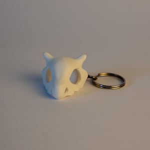 Può includere: Un portachiavi bianco con finitura opaca che presenta un design a teschio stilizzato con due piccole protuberanze a forma di corno sulla parte superiore, orbite oculari vuote e cavità nasale, fissato a un anello metallico argentato.