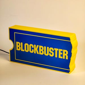 Può includere: Un'insegna rettangolare Blockbuster con un bordo giallo brillante e un centro blu intenso. La parola BLOCKBUSTER è stampata in grassetto con lettere maiuscole gialle al centro. L'insegna ha una forma a biglietto con lati dentellati e un cavo nero.