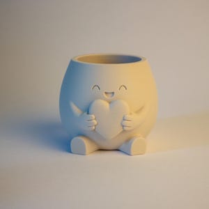 Può includere: Un vaso da fiori bianco sporco opaco a forma di personaggio arrotondato con un viso sorridente e gli occhi chiusi. La figura ha due braccia che tengono un cuore contro il petto e due piccoli piedi alla base. La parte superiore è aperta e circolare.